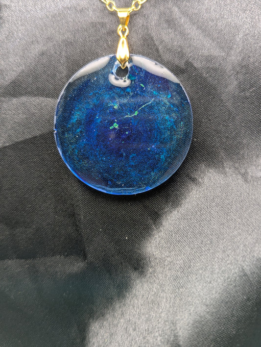 Blue resin circle pendant with speckles & swirls of green pigment powder. Resin necklace on a gold pinch bail and gold plated metal chain.