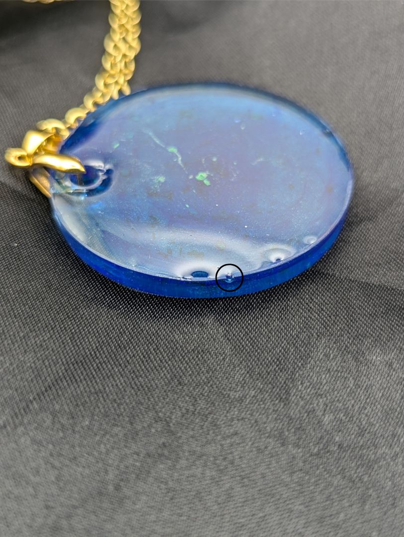 Blue resin circle pendant with speckles & swirls of green pigment powder. Resin necklace on a gold pinch bail and gold plated metal chain.