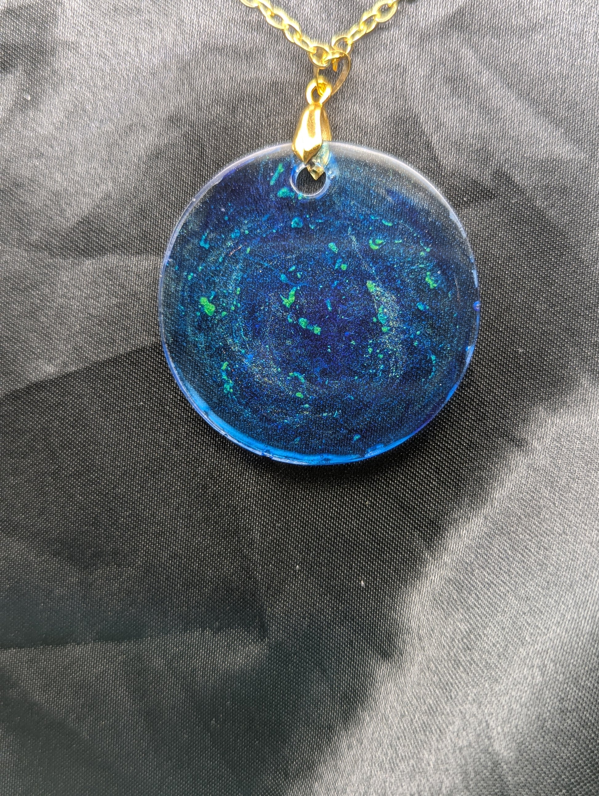 Back view of Blue resin circle pendant with speckles & swirls of green pigment powder. Resin necklace on a gold pinch bail and gold plated metal chain.