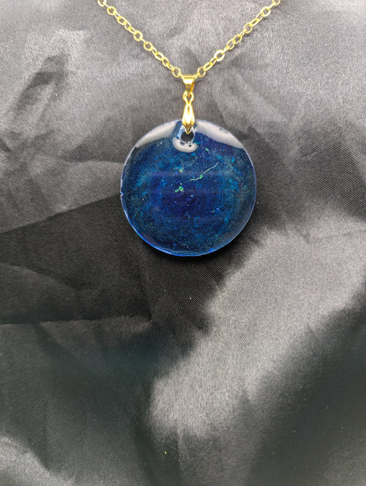 Blue resin circle pendant with speckles & swirls of green pigment powder. Resin necklace on a gold pinch bail and gold plated metal chain.