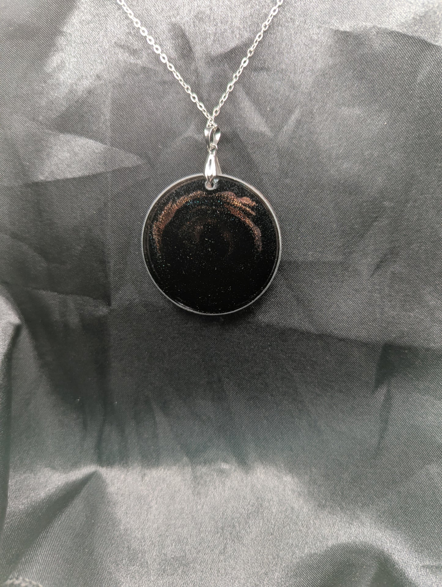Back view of Black Resin Circle Pendant with coral-pink colored sparkly swirls inside on a silver pinch bail & stainless steel chain.