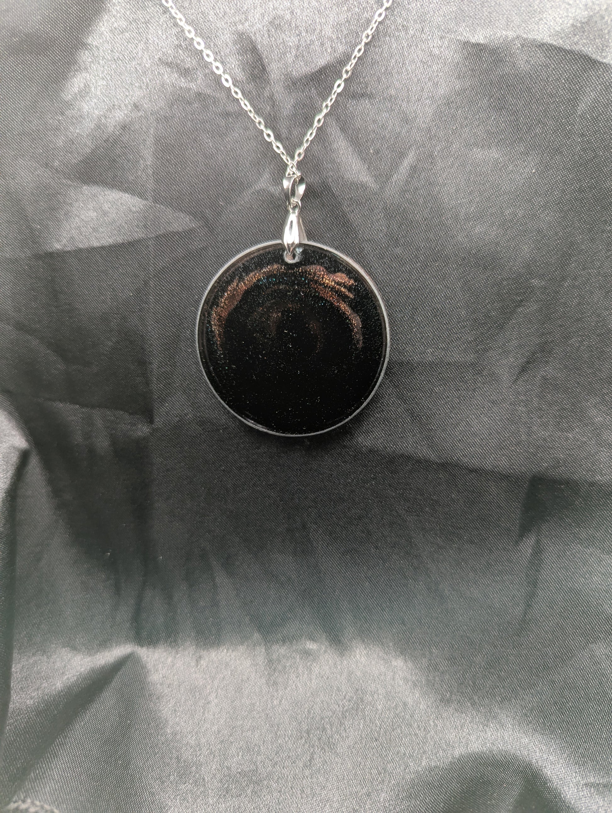 Back view of Black Resin Circle Pendant with coral-pink colored sparkly swirls inside on a silver pinch bail & stainless steel chain.