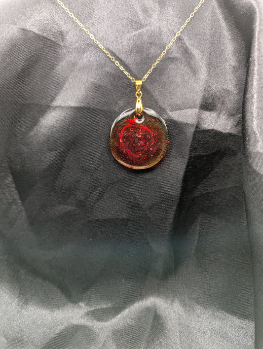 Red colored circle resin pendant with a coral colored pigment swirled in it, with a gold pinch bail on a gold plated metal chain.