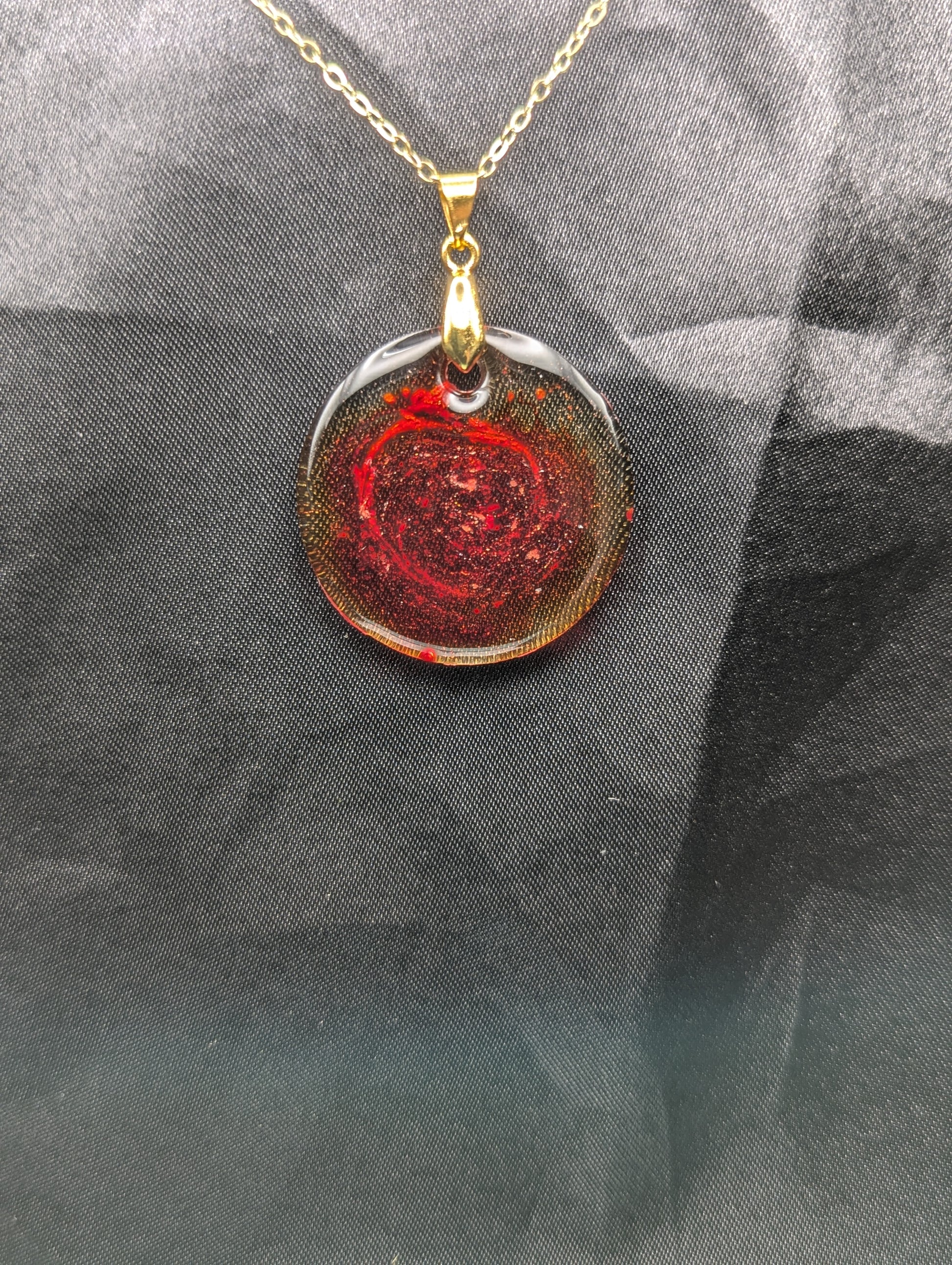 Red colored circle resin pendant with a coral colored pigment swirled in it, with a gold pinch bail on a gold plated metal chain.